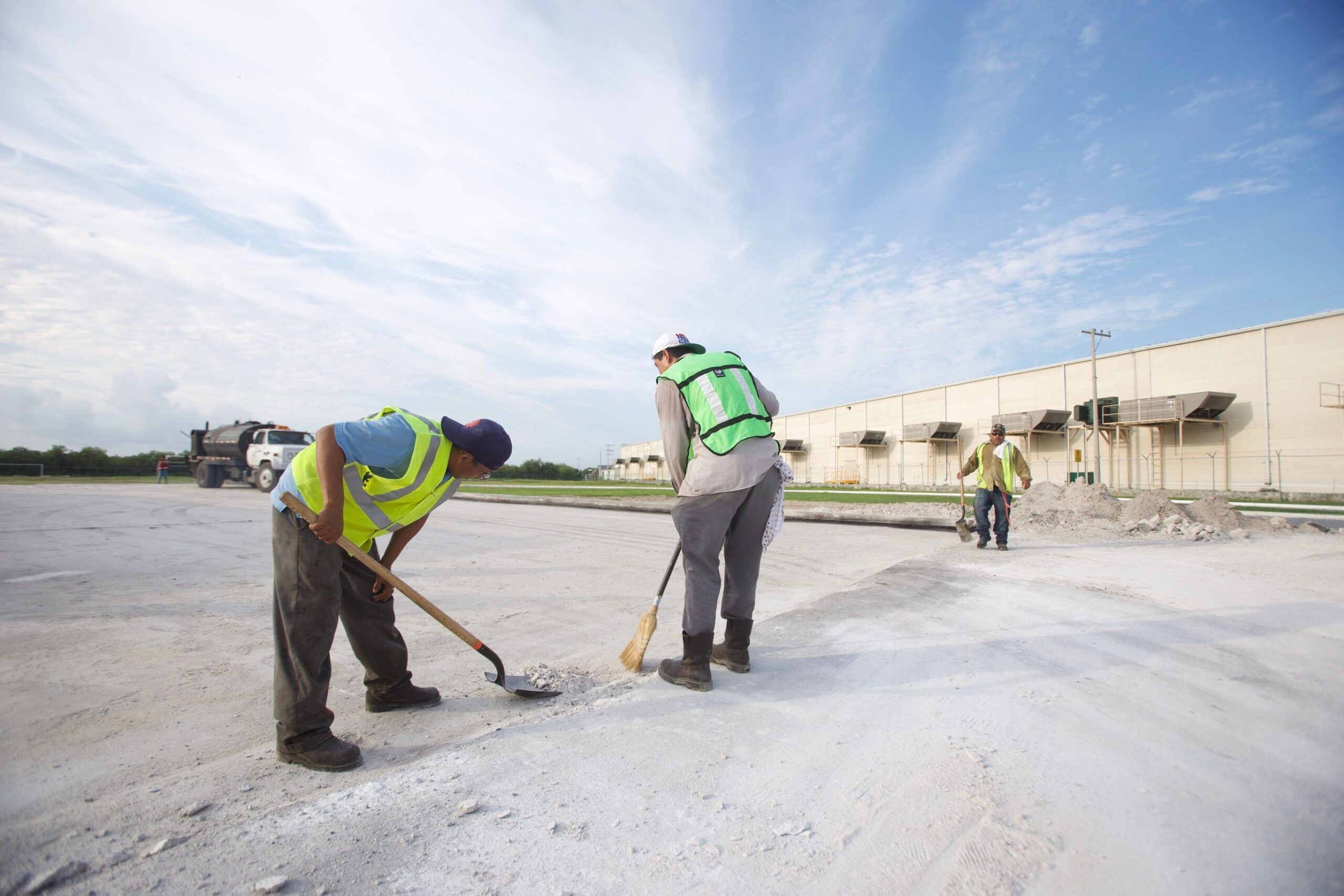 Workers in safety vests using brooms and shovels on a construction site under a bright sky.